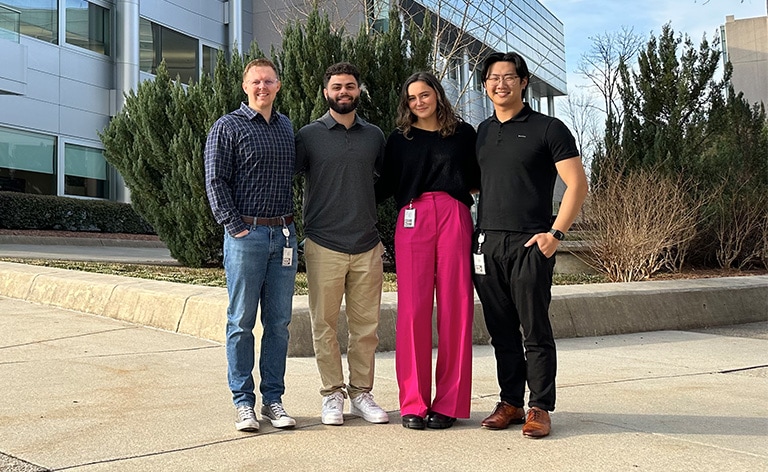 Four people stand together with a grey building in the background.