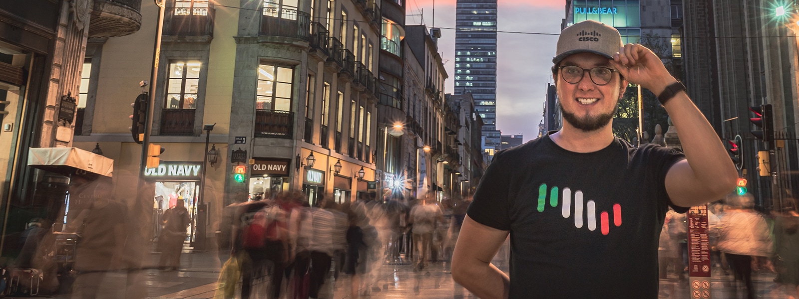 Male wearing a Cisco Hat and shirt holding left hand to his hat on a street in Mexico City